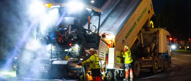 Workers guide an asphalt truck to pour in fresh asphalt into a paver