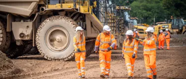 Four Aggregate Industries colleagues in high visibility workwear walk through Bardon Hill Quarry with heavy duty large dumper trucks in the background.