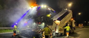 SuperCurve Asphalt being poured into a paver that's surfacing a road at night with workers around