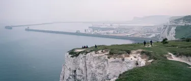 People walking and sitting on the edge of white cliffs with a large port seen in the distance