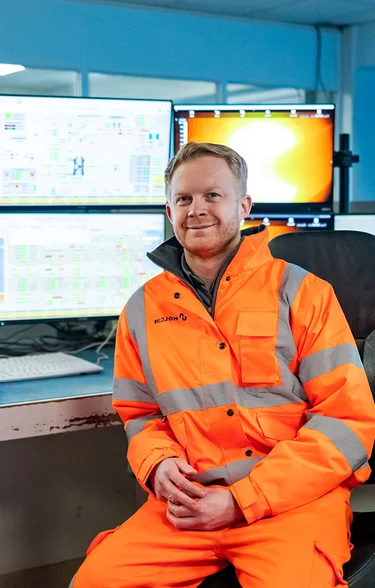 Man wearing PPE sitting in front of numerous computer monitors showing technical information