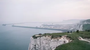 People walking and sitting on the edge of white cliffs with a large port seen in the distance