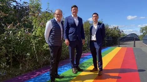Hr Director James Roberts, Surfacing Solutions Managing Director Tom Edgcumbe and High Viz LGBTQ+ Network Lead Aron Simpson-Webb stand on a new Pride themed pathway on the way into Leicester Pride at Abbey Park