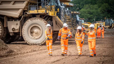 Four Aggregate Industries colleagues in high visibility workwear walk through Bardon Hill Quarry with heavy duty large dumper trucks in the background.