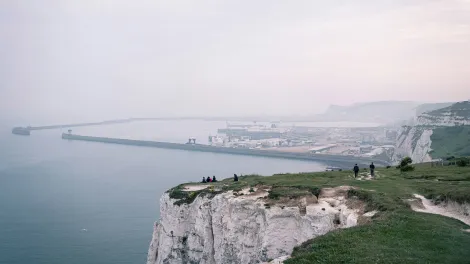 People walking and sitting on the edge of white cliffs with a large port seen in the distance