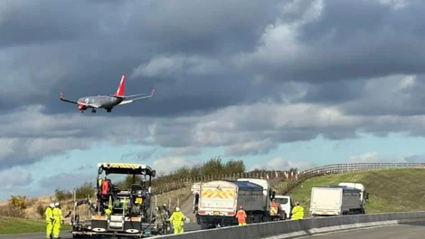 Teams ion high viz PPE carry out road resrtufacing works on the M42 Motorway Junction while an aeroplane flies overhead towards Birmingham Airport.