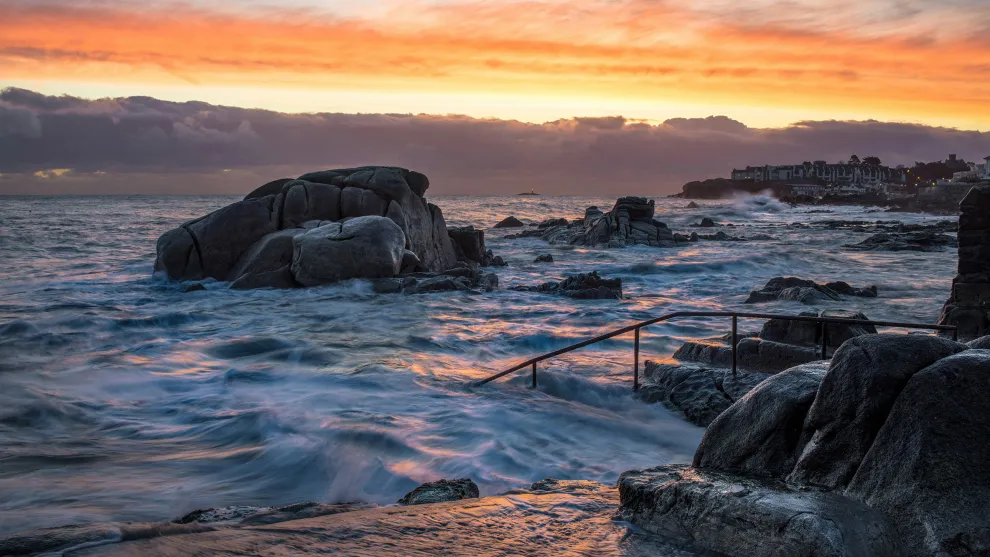 Sunset over a rocky coastal beach with a hand railing leading into the sea