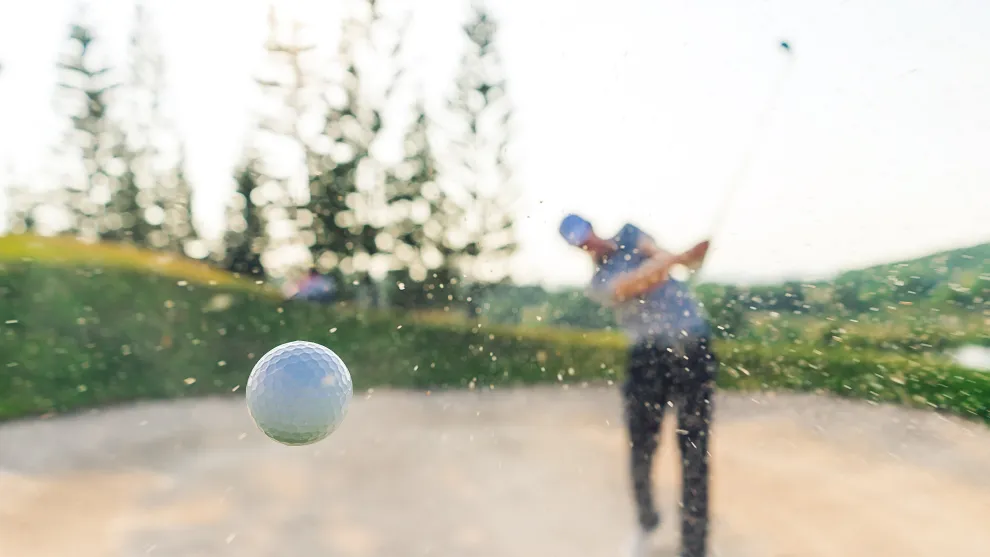 golf ball flying through the air with a golfer in a bunker behind