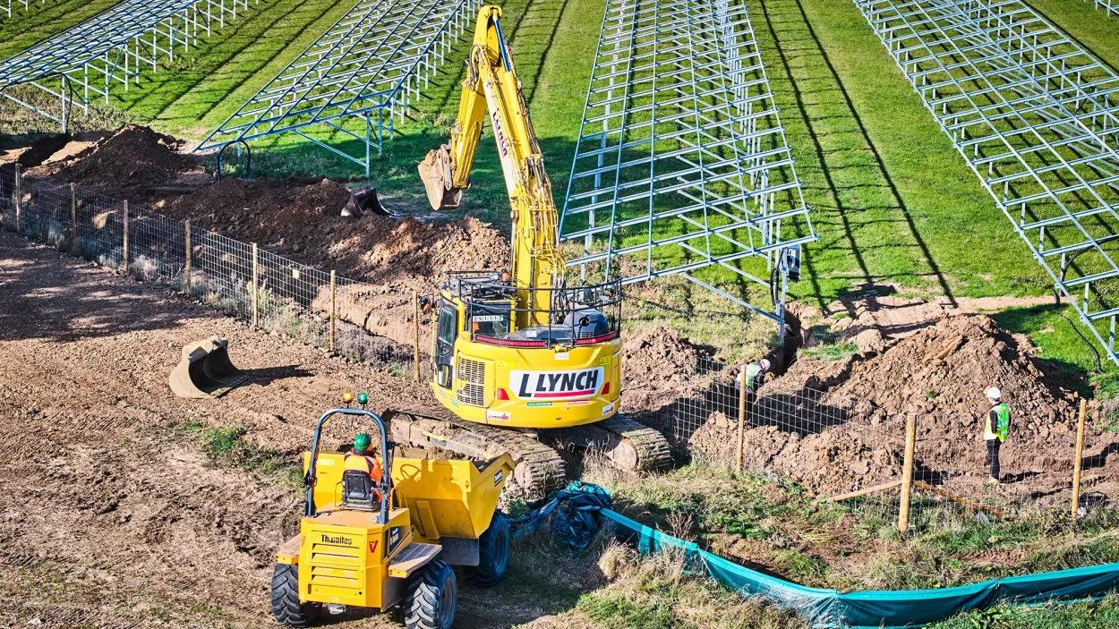 Excavator digging soil next to a loading shoval rows of solar panel frames