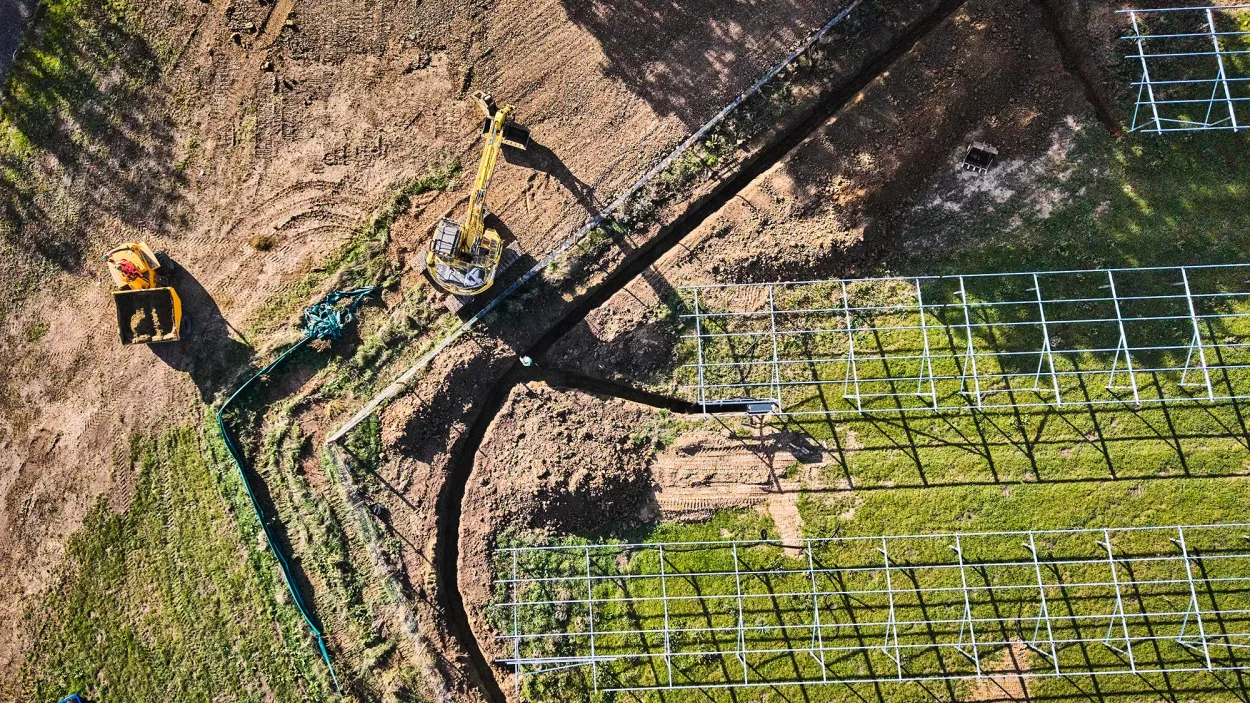 Top down image of a field with an excavator, loading shovel, trench and rows of solar panel frames