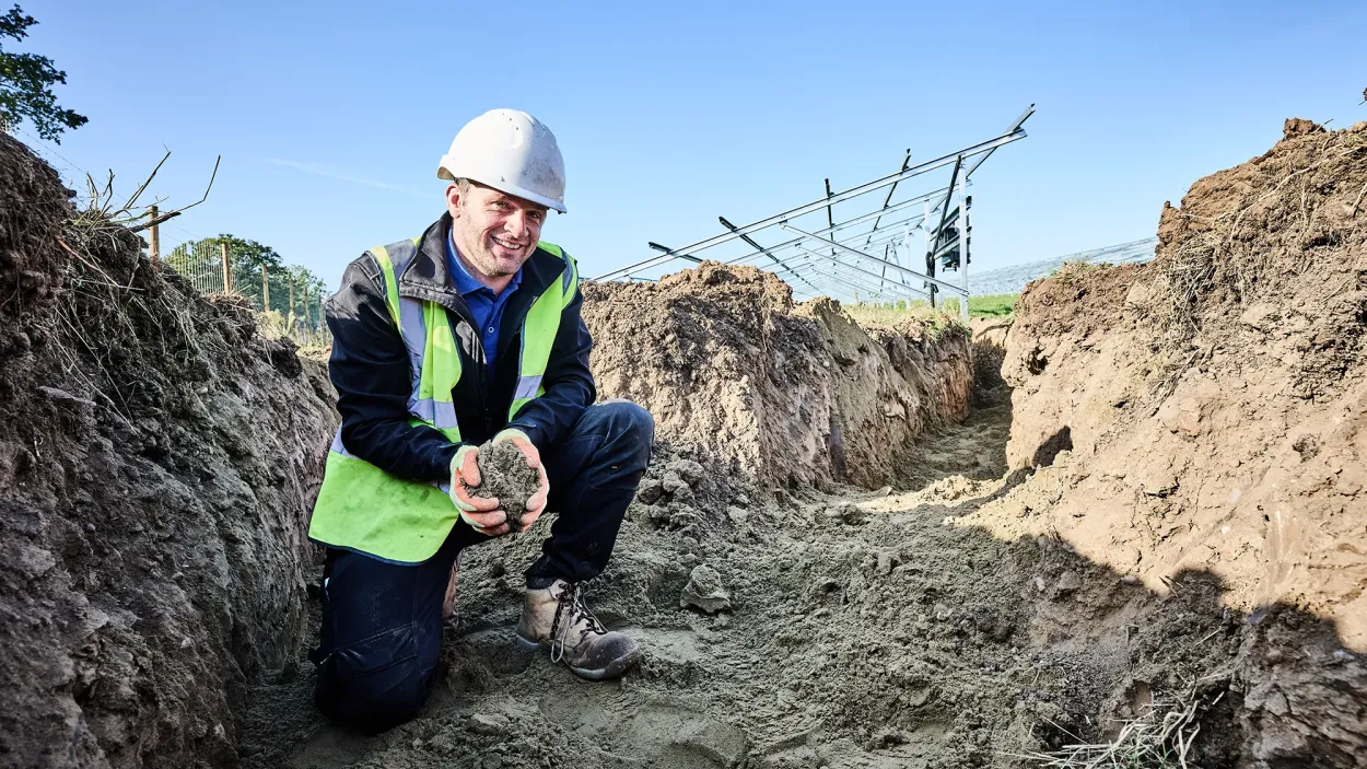 Man kneeling in a dirt trench holding soil with a solar panel frame behind him