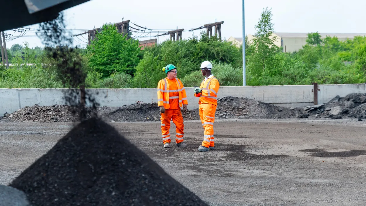 Mustapha Nyass, regional operation manager, at the Sheffield Asphalt Plant in high viz. He is standing in front of a pile of aggregates with a colleague.