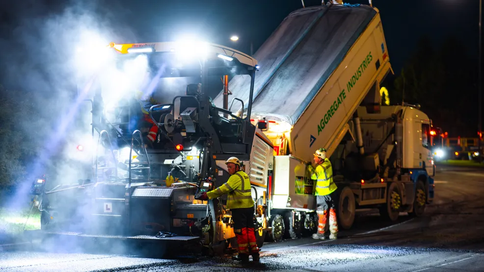 Workers guide an asphalt truck to pour in fresh asphalt into a paver