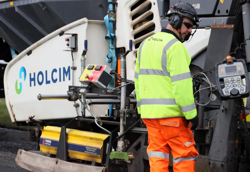 worker wearing PPE in front of an asphalt paver with the Holcim logo