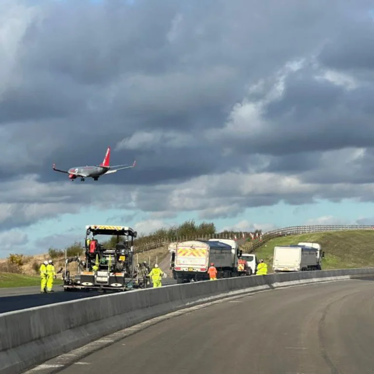 Teams ion high viz PPE carry out road resrtufacing works on the M42 Motorway Junction while an aeroplane flies overhead towards Birmingham Airport.