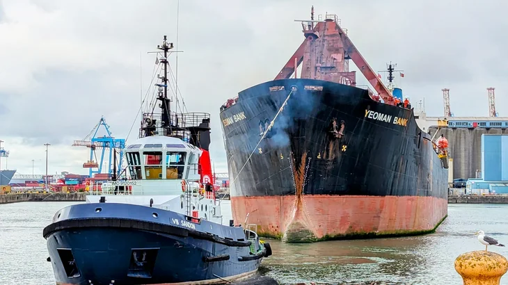 The Yeoman Bank ship being pulled through a harbour by tug boat
