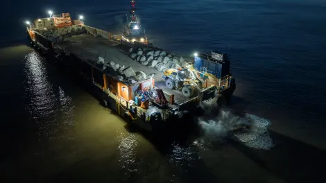 A sea barge at night carries a delivery of Armourstone granite boulders from Glensanda to Blue Anchor Beach, Somerset.