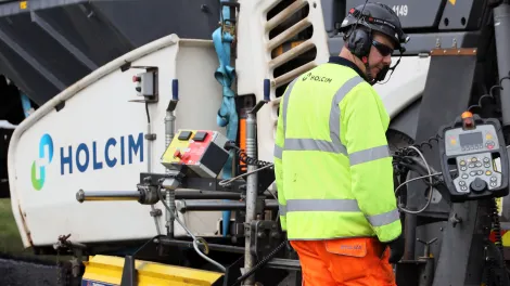 worker wearing PPE in front of an asphalt paver with the Holcim logo