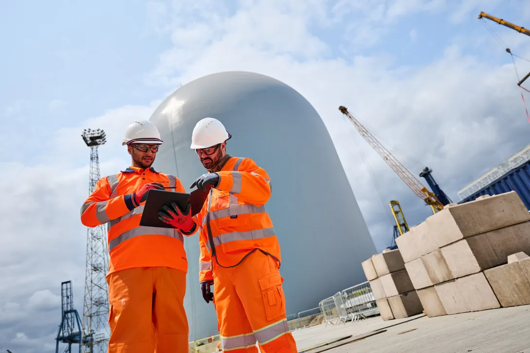 Two Holcim colleagues dressed in high viz PPE reviewing plans at the Holcim UK Tilbury Cement Terminal with the dome silo in the background.