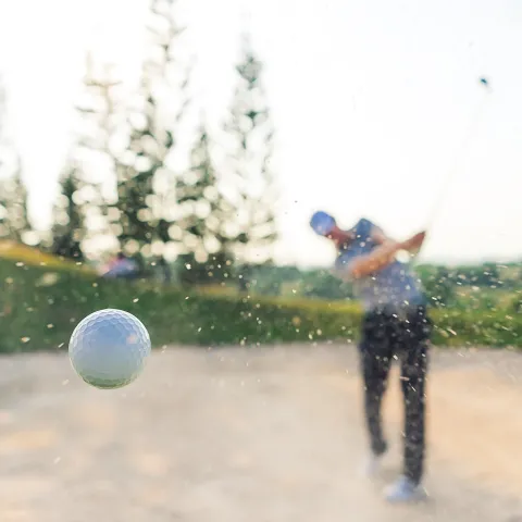 golf ball flying through the air with a golfer in a bunker behind
